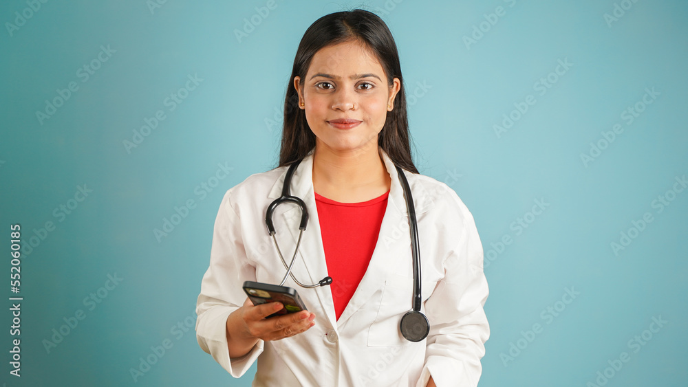 Portrait of a young female doctor holding mobile phone in her hand, Asian Indian woman doctor in apron isolated over blue studio background, Using smart phone Portrait of a young female doctor holding mobile phone in her hand, Asian Indian woman doctor in apron isolated over blue studio background, Using smart phone