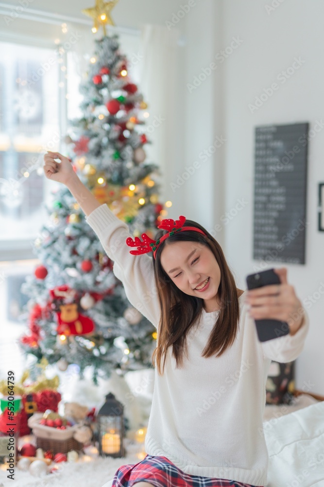 Cute beautiful young asian lady woman wearing reindeer headband selfie and video call with mobile phone posing in front a big Christmas tree with lots of decoration lights gift box and ornaments Cute beautiful young asian lady woman wearing reindeer headband selfie and video call with mobile phone posing in front a big Christmas tree with lots of decoration lights gift box and ornaments