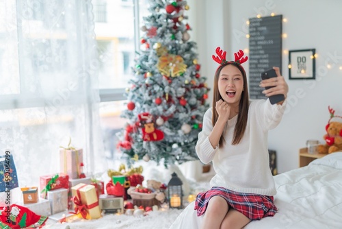 Cute beautiful young asian lady woman wearing reindeer headband selfie and video call with mobile phone posing in front a big Christmas tree with lots of decoration lights gift box and ornaments © asean studio
