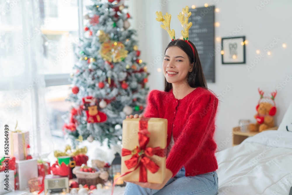 Cute beautiful young asian lady woman wearing reindeer headband holding a gold color gift box with red ribbon posing in front a big Christmas tree with lots of decoration lights gift box and ornaments Cute beautiful young asian lady woman wearing reindeer headband holding a gold color gift box with red ribbon posing in front a big Christmas tree with lots of decoration lights gift box and ornaments