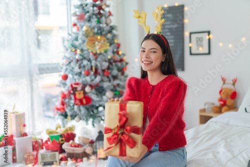 Cute beautiful young asian lady woman wearing reindeer headband holding a gold color gift box with red ribbon posing in front a big Christmas tree with lots of decoration lights gift box and ornaments © asean studio