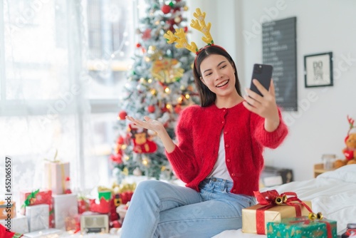 Christmas celebration. Young asian lady woman wearing reindeer headband selfie and video call with mobile phone posing in front a Christmas tree with lots of decoration lights gift box and ornaments