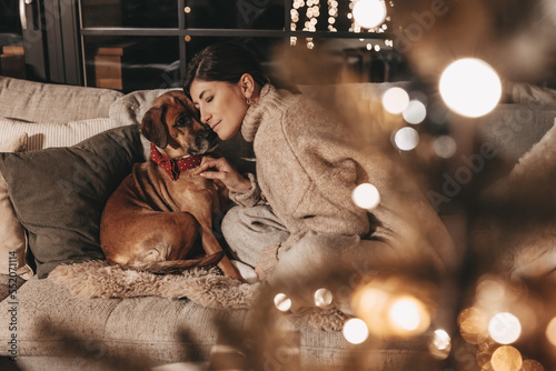 Happy woman with dog cuddling and unpacking gifts, looking excitedly into the box, in a cozy environment at home on the couch, mysterious, warm colors