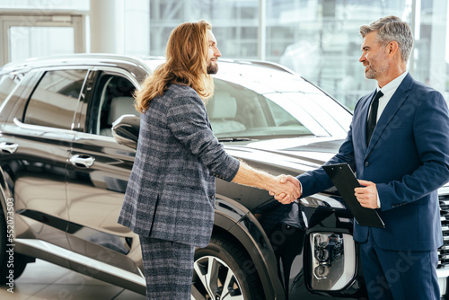 Happy middle age car salesperson came to an agreement with young long haired beard man in a showroom with new crossover on background. © Iryna