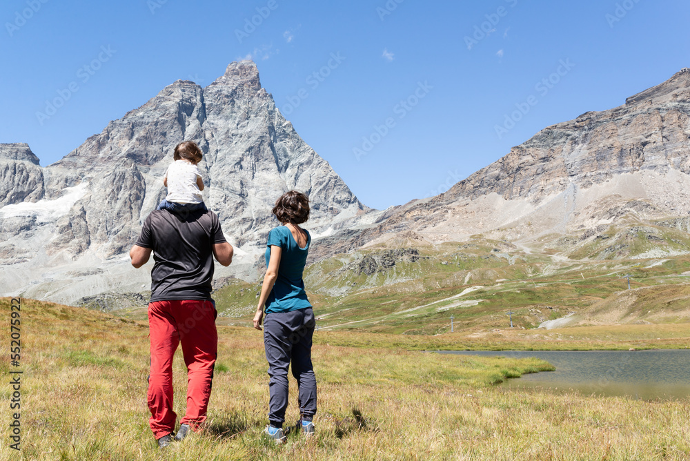 Rear view of a standing mother and father with his little daughter on shoulders looking at Matterhorn during a hiking day in Cervinia, italian alps. 