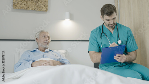 Doctor talking to senior patient in bed, writing down in patient's card, elderly care