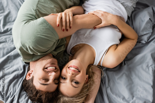 Top view of cheerful young couple in pajama looking at camera while hugging on bed.