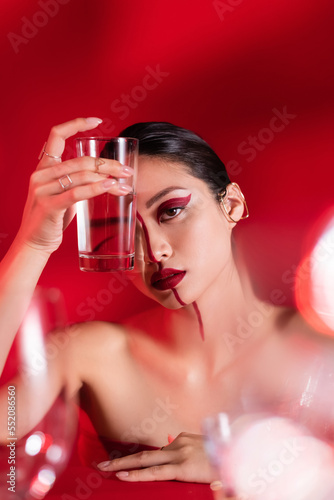 brunette woman with makeup and bare shoulders holding glass of water near face on red background.