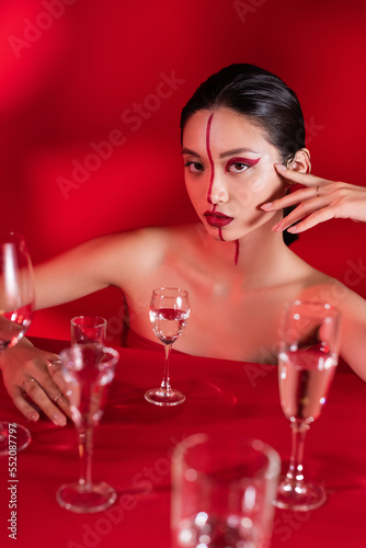 asian woman with bright creative visage and hand near face looking at camera near various glasses on red background.