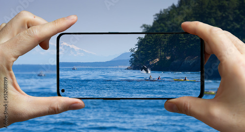 Tourist taking a picture with a mobile phone at a whale wathing tour while a Orca is jumping out of the water.