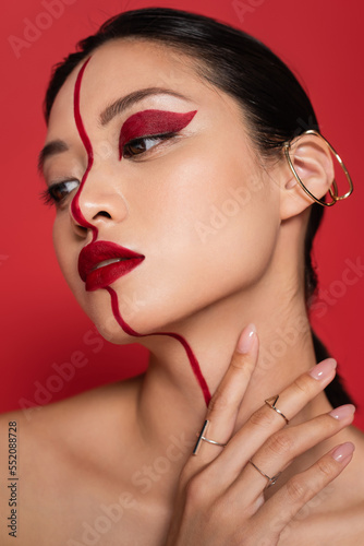 portrait of brunette asian woman with creative makeup and cuff earring holding hand near neck isolated on red.