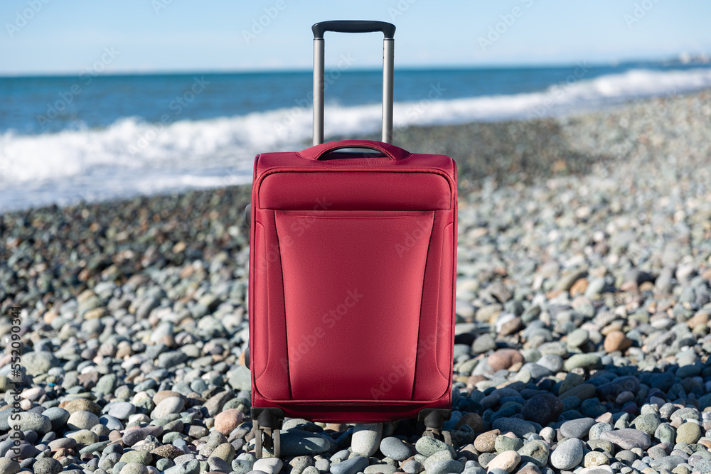Color travel suitcase on pebble beach with turquoise sea background, summer holidays concept
