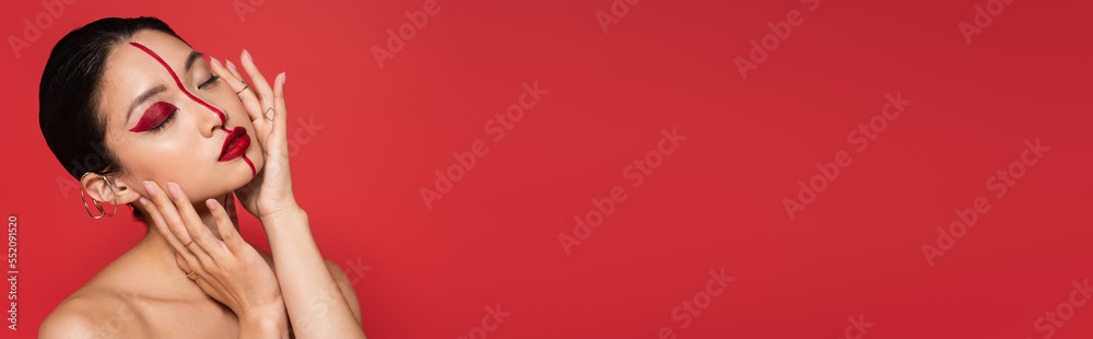 Fototapeta premium sensual asian woman with bare shoulders and artistic visage touching perfect face with closed eyes isolated on red, banner.