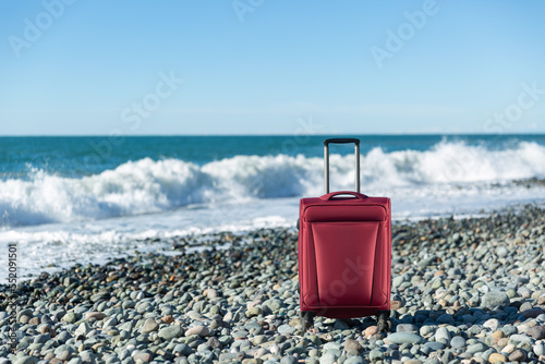 Color travel suitcase on pebble beach with turquoise sea background, summer holidays concept