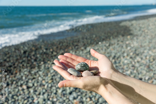 female hands holding small pebble stones in hands near blue sea on a beach background, picking up pebbles on the stone beach, round shape pebbles, summer vacation souvenir, beach day