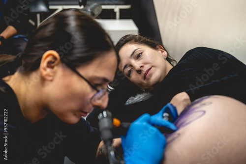 Concentrated young tattoo artist girl with glasses making a tattoo, with tattoo machine, of a big tiger, in the thigh and hips of a happy and beautiful woman
