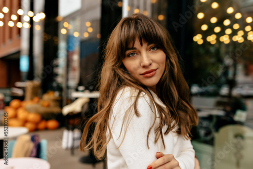 Stylish pretty girl with dark wavy hair wearing knitted white sweater posing at camera on city lights. She is smiling and crossed arms on waist