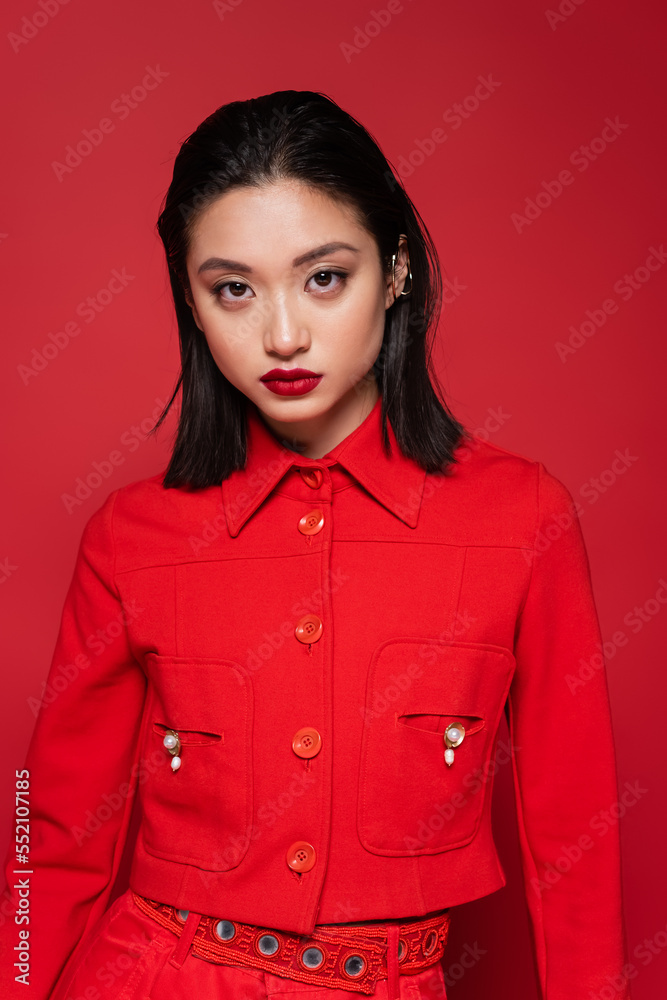 brunette asian woman with makeup wearing stylish jacket and looking at camera isolated on red. brunette asian woman with makeup wearing stylish jacket and looking at camera isolated on red.
