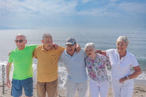 gruppo di 5 anziani al mare si abbracciano felici - sullo sfondo si vede il cielo blu