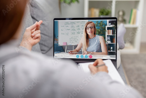 Back view of mature woman on comfy couch listening online lesson of wireless laptop. Lady studying english in light living room interior