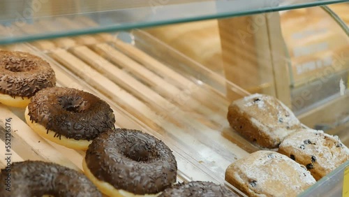 A customer picks up sweet pastries, cupcakes, and donuts from a display case in a bakery using serving tongs. Baking and flour food threatens to increase excess weight.