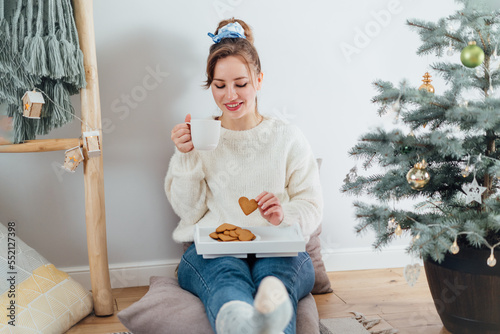 Happy Young Woman in cosy sweater drinking hot tea with cookies, relaxing on floor cusions near potted christmas tree in modern Scandi interior home. Eco-friendly cozy winter holidays. Selective focus photo