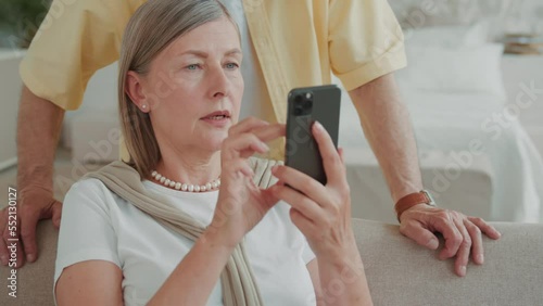 Close up elder woman use smartphone relaxing on sofa at home husband man standing on background smiling feel happy. Choose discussing. Have fun enjoy mobile application. Modern gadget. Communication