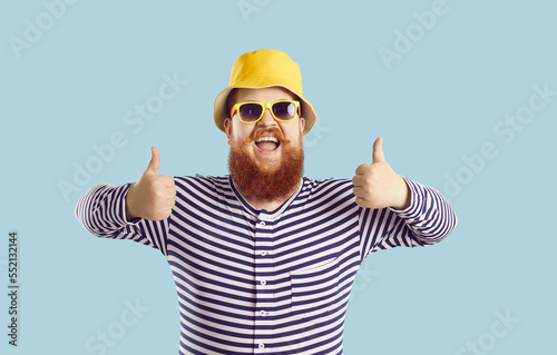 Portrait of cheerful funny chubby man showing thumbs up isolated on pastel light blue background. Red-bearded man in panama, sunglasses and striped clothes with funny expression recommends something.
