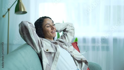 Beautiful dark haired young lady relaxing on comfortable sofa with hands behind head in the light living room Smiling peaceful curly girl enjoy calm leisure time at home alone No stress people concept photo