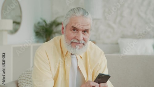 Senior man uses a smartphone, browses and reads share text messages. Sitting on sofa at home looks at screen, swipes. Portrait. Close up view.