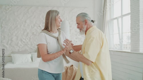 Happy old couple dancing in the living room, the loving husband holds the hand of his beloved wife. Enjoy life together and feel happy. Modern apartments