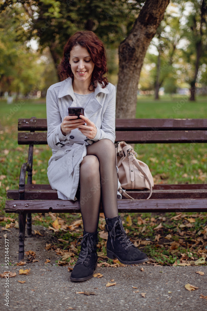 Pretty laughing girl with smartphone has a good time in autumn weekend. Outdoor portrait of lovable trendy lady wear grey coat in october day. Pretty laughing girl with smartphone has a good time in autumn weekend. Outdoor portrait of lovable trendy lady wear grey coat in october day.