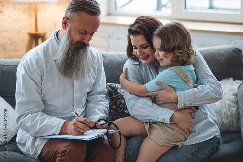 Kind male pediatrician doctor visiting his patient at home, examining little girl sitting on mother's lap, writing prescription. Concept of kid's health check