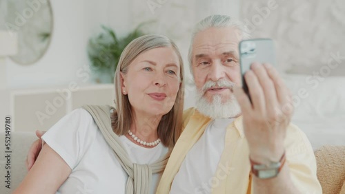 Portrait of happy elderly couple chatting online with their relatives and children waving hands sitting on sofa at home. Smiling. Modern apartment. Communication