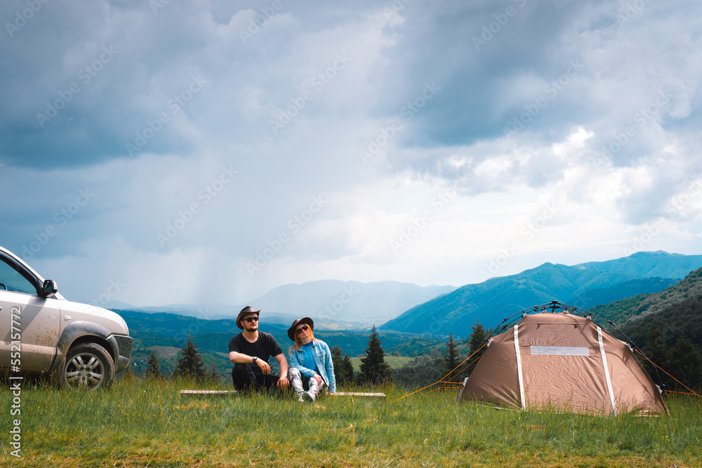 On the top of the mountain, a couple, a man and a woman, are enjoying the view of the mountains. A tourist tent and a four-wheel drive car with a roof rack. concept of freedom and travel, Ukraine