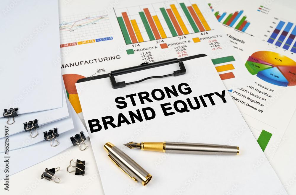 On the table among financial graphs and charts lies a tablet with the inscription - STRONG BRAND EQUITY On the table among financial graphs and charts lies a tablet with the inscription - STRONG BRAND EQUITY