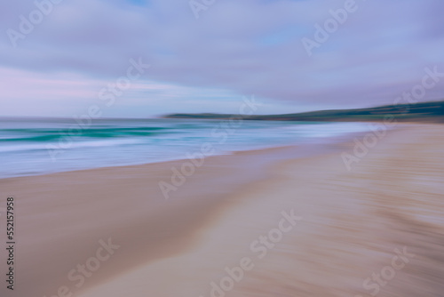 Abstract seascape. Wide sandy beach, turquoise water, cloudy sky, and mountains. Motion blur, light blue, and pink colors