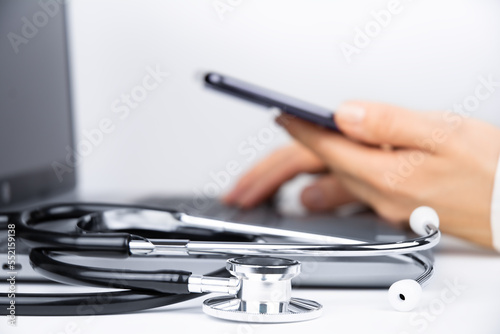 Female doctor's hands working on laptop with smartphone, stethoscope on foreground. Telemedicine concept