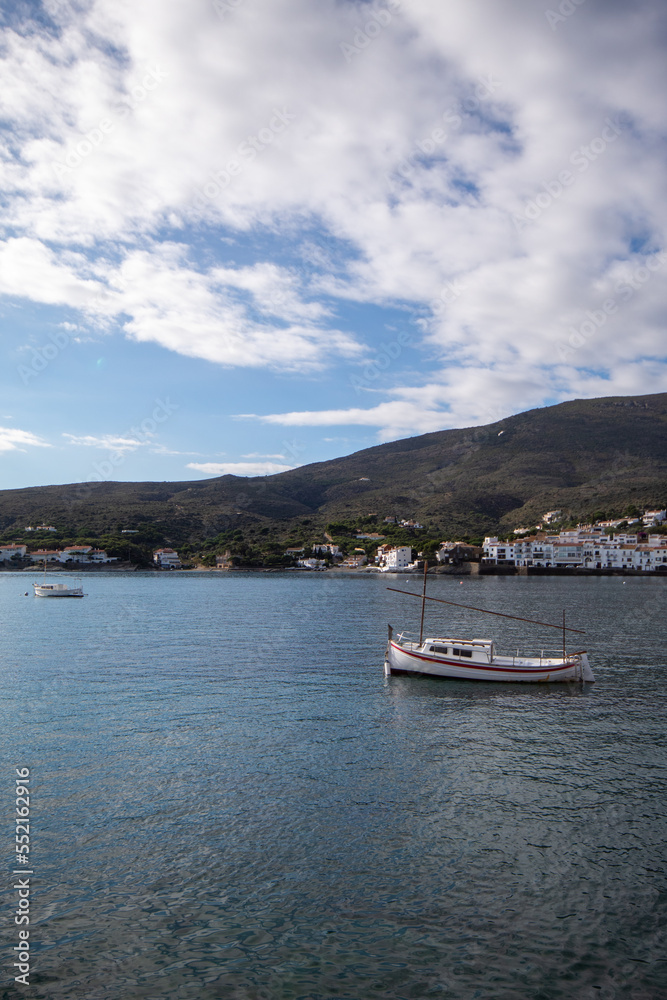 Barco blanco en medio del mar del pueblo de Cadaqués con el pueblo y la verde montaña de fondo bajo un cielo azul con las blancas nubes.