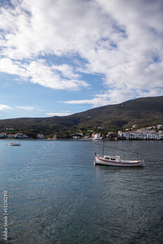 Barco blanco en medio del mar del pueblo de Cadaqu  s con el pueblo y la verde monta  a de fondo bajo un cielo azul con las blancas nubes.