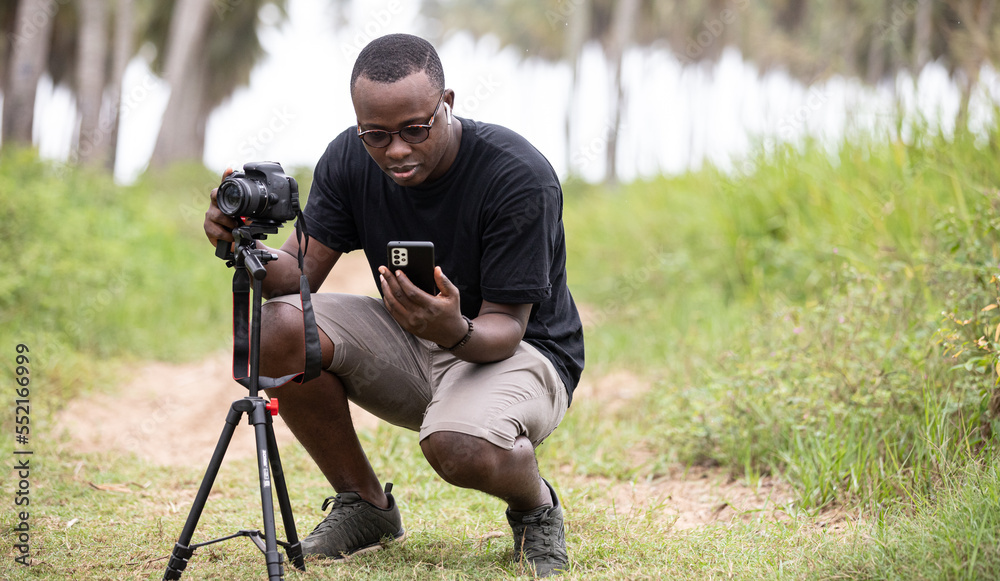 Portrait of an African photographer taking a picture with a digital camera while setting up his mobile phone for remote control