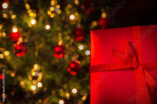 Uma caixa de presente, vermelha, com um laço de fita, com uma árvore de Natal ao fundo. photo