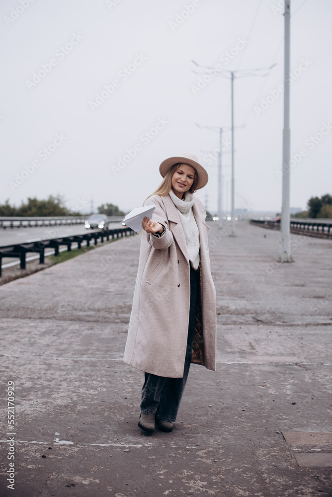 Portrait of pretty girl hold origami plane. Woman in felt hat and gray coat and sweater with paper airplane outdoor in autumn