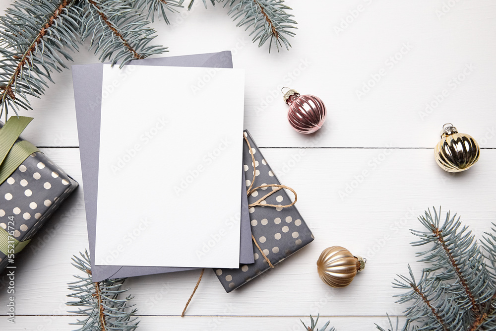 Fototapeta premium Christmas greeting card mockup with grey envelope, gift boxes and decorations on white wooden background, top view, flat lay. Winter holiday card mockup with fir tree branches and balls