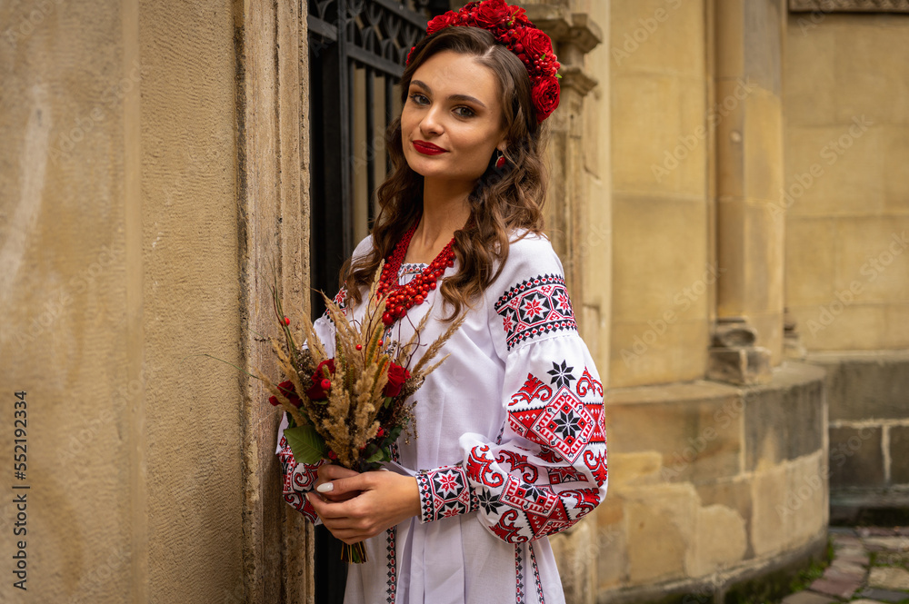 Fototapeta premium Portrait of a beautiful Slavic girl with long blond hair and blue eyes with a flower crown in a white and red embroidered dress with a bouquet. Traditional clothes of the Ukrainian region