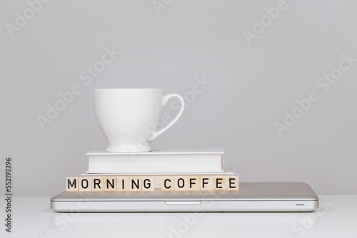 White porcelain mug on the white minimalistic table with books  laptop and wooden letters that make an inscription - morning coffee on grey background 