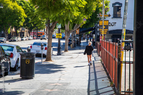 a woman wearing white headphones, a black hoodie, and short walking along a sidewalk in Hollywood with cars parked and driving and lush green trees in Los Angeles California USA