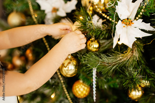 Child s hands decorating Christmas tree with shiny gold ball. The tree is decorated with xmas silver and gold baubles  garlands and lights. Christmas and new year family holiday celebration concept