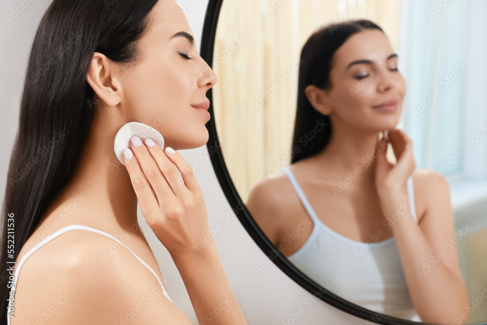 Young woman using cotton pad with micellar water near mirror indoors Young woman using cotton pad with micellar water near mirror indoors