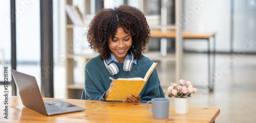Cute african american female student wearing headphones with afro dreadlocks, studying remotely from home, using a laptop, taking notes on notepad during online lesson, e-learning concept, 